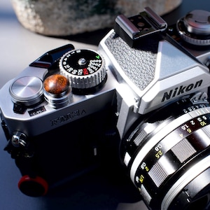 May include: A silver and black Nikon film camera featuring a textured grip, a prominent shutter speed dial with white and red markings, a circular brown shutter release button, and a black lens with silver focus rings and yellow distance indicators.