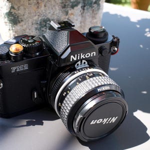 May include: A black Nikon FM2 35mm film camera featuring a silver and black lens with a Nikon lens cap. The camera body includes a textured grip, a shutter speed dial with white numbers, and a small yellow circular button on the top right side.