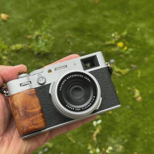 May include: A silver and black digital camera featuring a textured grip, a wooden handle attachment, and a lens labeled FUJINON ASPHERICAL LENS SUPER EBC f=23mm 1:2.8. The camera body includes a top-mounted shutter button and a viewfinder window.