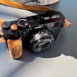 May include: A black Fujifilm X100VI digital camera featuring a custom light brown wooden hand grip and a matching wooden shutter release button. The camera includes a tan leather neck strap and a lens with white text reading FUJIFILM ASPHERICAL LENS.