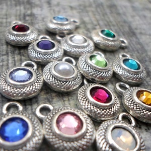 May include: Assortment of silver-toned charms with various colored rhinestones. Each charm features a round, faceted rhinestone in colors such as red, blue, green, purple, pink, and white. The charms have a decorative, textured design.