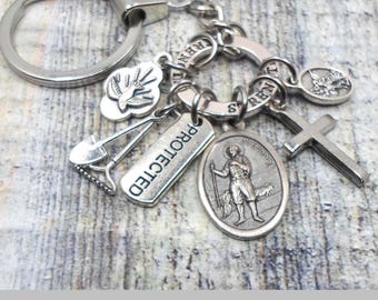Può includere: Portachiavi argentato con diversi ciondoli, tra cui una croce, una colomba e un ciondolo con la scritta "PROTECTED". Il portachiavi presenta anche un ciondolo con una figura di santo. Il testo "Necklace, Keychain or Clip" è in basso.