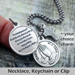 May include: Silver necklace with a charm that reads "Remind yourself that it's not about getting through the week or month or year, but getting by one day at a time..." and another charm with a pair of hands clasped in prayer. The necklace also has a Celtic cross charm.
