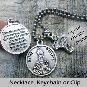 May include: Silver necklace with a Saint Patrick charm and a Celtic cross charm. The necklace has a ball chain and the charms are attached to a ring. The Saint Patrick charm has an image of Saint Patrick and the text "Saint Patrick". The Celtic cross charm has a Celtic knot design. The necklace is available as a necklace, keychain, or clip. The text "May the road rise to meet you, May the wind be always at your back. May God hold you the palm of His Hand" is on the back of the Saint Patrick charm.