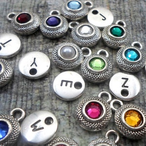 May include: Assortment of silver charms with various colored rhinestones and letters. The charms have a textured surface and a hole for attaching to a chain or bracelet.