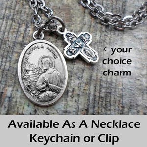 May include: A silver-toned necklace with two charms. One charm is an oval pendant with the text "S. Angela Merici" and a depiction of a saint. The other charm is a cross. The text "Available As A Necklace Keychain or Clip" is at the bottom.