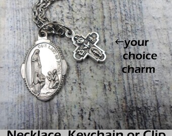 Può includere: Una collana color argento con una catena e due ciondoli. Uno è un ciondolo ovale con l'immagine di Nostra Signora di Fatima e due figure in ginocchio. L'altro è un ciondolo a forma di croce. Il testo in basso dice "Necklace, Keychain or Clip."