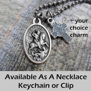 May include: A silver-toned necklace with two charms. One charm is an oval pendant depicting St. George on horseback slaying a dragon. The other is a cross-shaped charm. The text "Available As A Necklace Keychain or Clip" is at the bottom.