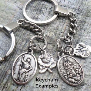 Pu&ograve; includere: Portachiavi d'argento con quattro ciondoli: una rosa, una colomba e due medaglioni ovali con immagini religiose. I medaglioni presentano il testo "St Joan Arc" e una rappresentazione di un pastore con pecore. Il testo "Keychain Examples" &egrave; visibile sotto i ciondoli.