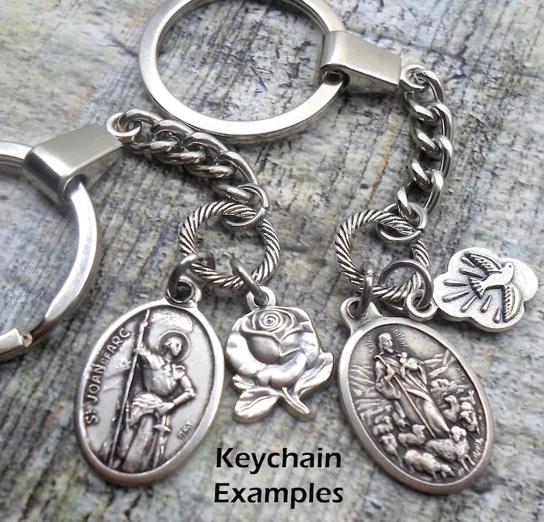 Pu&ograve; includere: Portachiavi d'argento con quattro ciondoli: una rosa, una colomba e due medaglioni ovali con immagini religiose. I medaglioni presentano il testo "St Joan Arc" e una rappresentazione di un pastore con pecore. Il testo "Keychain Examples" &egrave; visibile sotto i ciondoli.