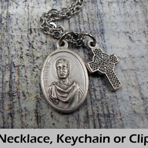 May include: Silver-toned necklace with an oval pendant featuring a religious figure and a Celtic cross charm. The chain is a linked design. The text at the bottom reads "Necklace, Keychain or Clip."
