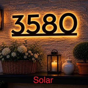 Peut inclure: Panneau de numéro de maison éclairé 3580 avec une lueur chaleureuse, monté sur un mur texturé. Le mot "Solar" est visible en bas. La scène comprend un panier de fleurs, une lanterne et une plante en pot.