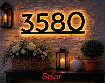 May include: Illuminated address numbers 3580 with a warm glow, mounted on a textured wall. The word "Solar" is visible at the bottom. The scene includes a basket of flowers, a lantern, and a potted plant.