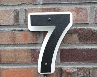 May include: A white and black house number sign with the number 7 in black on a white background. The sign is mounted on a brick wall.