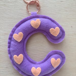 May include: A purple felt letter "C" keyring with peach-coloured heart accents. The keyring has a rose gold clasp. The felt letter is a soft, plush material.