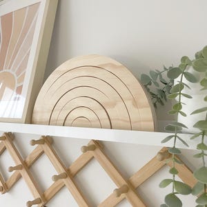 Puede incluir: Un juguete de apilamiento de arcoíris de madera con múltiples arcos se encuentra en un estante blanco. A la izquierda hay una impresión enmarcada con un diseño de sol. Debajo del estante hay un perchero de madera extensible. Una planta con hojas verdes está a la derecha.
