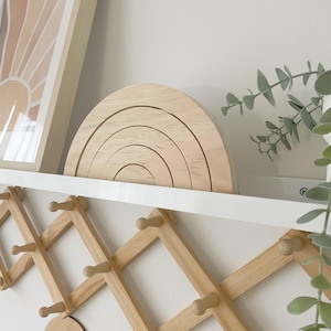 May include: A wooden rainbow arch with five rings stacked on top of each other. The arch is on a white shelf with a wooden coat rack underneath. The coat rack has a diamond pattern and wooden knobs. The text "COTTON FOX" is on a round wooden tag hanging from the rack.
