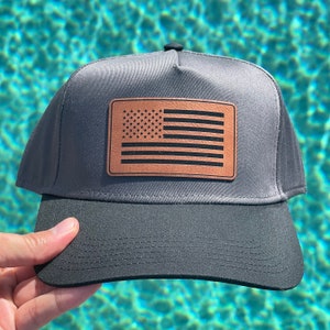 May include: A black and grey baseball cap with a brown leather patch featuring a black and white American flag.