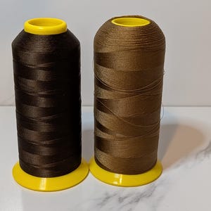 May include: Two spools of brown thread. One spool is a dark chocolate color, and the other is a lighter, caramel brown. Both spools have a yellow base and top. The thread is tightly wound, and the spools are set against a neutral background.