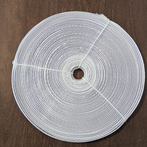May include: A tightly wound roll of white ribbon, secured with a thin, light green string. The ribbon is neatly coiled, revealing a series of concentric circles. The background is a dark brown wooden surface.