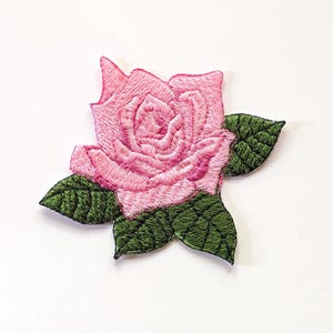 May include: Embroidered patch of a pink rose with green leaves. The rose is in full bloom, showcasing detailed stitching and varying shades of pink. The leaves are a deep green, providing contrast to the rose.