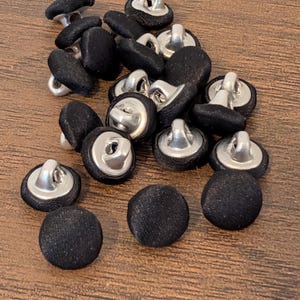 May include: A collection of black fabric-covered buttons with silver metal shanks. The buttons are round and have a slightly textured appearance. They are scattered on a wooden surface.