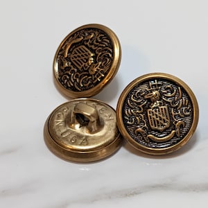 May include: Three antique brass buttons with a detailed crest design. The buttons have a raised emblem with ornate scrollwork and a shield. One button has the text "USA" on the back.