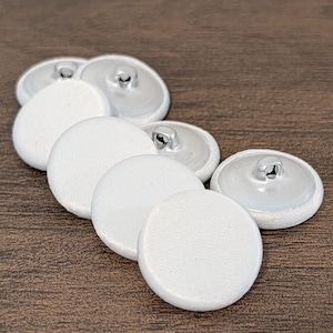 May include: A collection of white fabric-covered buttons with silver metal backs. The buttons are round and arranged on a dark brown wooden surface. The fabric has a subtle texture.