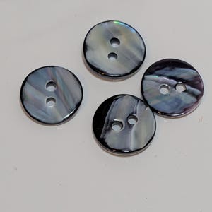 May include: Four round buttons with two holes each. The buttons have a pearlescent, iridescent finish with shades of black, gray, and white. The buttons are arranged on a white surface.