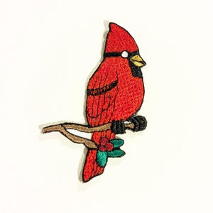 May include: Embroidered patch of a red cardinal bird perched on a brown branch. The bird has black accents, a yellow beak, and a white spot. Green and red leaves and berries are at the base of the branch.