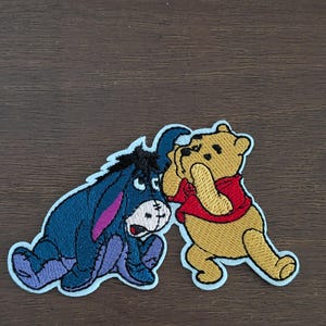 May include: Embroidered patches of Winnie the Pooh and Eeyore. Winnie the Pooh wears a red shirt and has his hands up. Eeyore is blue with a pink inner ear and purple hooves. The patches have a white border.