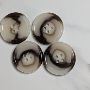 May include: Four round buttons with a swirl pattern in shades of brown and cream. Each button has four holes for sewing. The buttons are made of a glossy material.