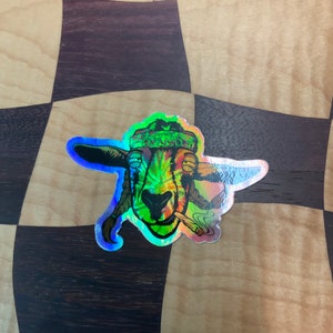 May include: A holographic sticker of a goat wearing a bandana with the word "Blessed" on it. The goat is smoking a joint and has a rainbow-colored design.