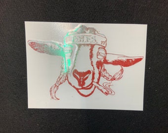 May include: A white sticker featuring a red illustration of a goat's head with braided hair and a cigarette. The goat wears a headband with the text "B.M.F.S. 73". The image has a holographic effect.