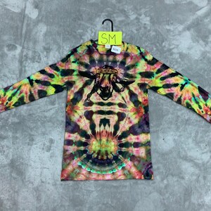 May include: A long-sleeved tie-dye shirt with a colorful psychedelic pattern. The shirt features a black and white graphic of a goat wearing a bandana and smoking a pipe. The text "PAISLEY" is written on the bandana. The shirt is labeled "SM".