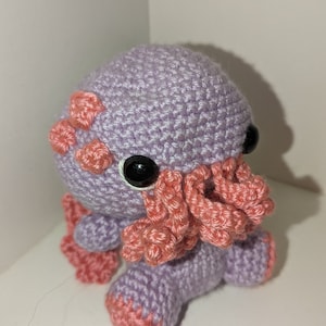 May include: A handmade crochet plush toy in the shape of a purple octopus with pink accents. The octopus has large black button eyes and pink tentacles. The crochet stitches are visible, giving the toy a textured appearance.
