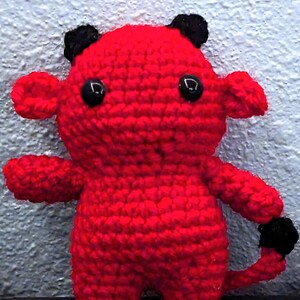 Puede incluir: Un juguete de peluche rojo tejido a crochet con cuernos negros y cola. El juguete tiene grandes ojos negros y una expresión simple y amigable.