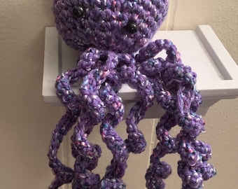 May include: A crocheted purple octopus with black eyes. The octopus has a speckled purple body and eight tentacles.