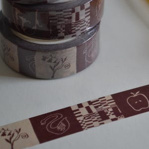 Könnte beinhalten: Dekorative Washi-Tape-Rollen mit einem sich wiederholenden Muster aus braunen und beigen Quadraten, Schwänen, Äpfeln und Sternen. Das Klebeband ist teilweise abgerollt und zeigt das Design. Die Rollen sind auf einer weißen Oberfläche gestapelt.