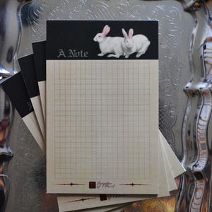 May include: A stack of rectangular memo pads with a cream-colored grid paper body. The top section is black with two white rabbits and the text A Note. The bottom features a small red square logo and the text Daughter of Pearl.