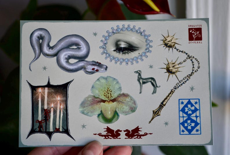 May include: A postcard featuring various illustrations, including a silver snake, an eye surrounded by pearls, a dog, a flower, candles, and a chain with spiky balls. The postcard has a light blue border and the text "DAUGHTER OF PEARLS".