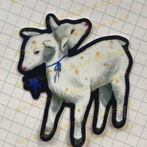 May include: A sticker depicting a two-headed lamb illustration. The lamb is mainly white with grey shading and pink details. It has a blue ribbon and a blue star graphic. The sticker has a black border and a grid background.