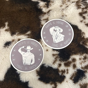 May include: Two round stickers with a cowboy theme. Each sticker features a cowboy illustration in white on a gray background, surrounded by stars. The text "SAVE A HORSE RIDE A COWBOY" is printed on each sticker.