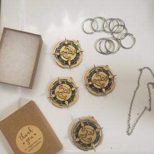 Op de afbeelding: Vier gouden en groene decoratieve bedels met een draaiend ontwerp, een ketting en een set zilveren sleutelringen. De bedels hebben een groene kern met een gouden draaiend ontwerp en een gouden rand. De bedels liggen op een wit oppervlak. Een bruine doos met de tekst "Bedankt voor het steunen van ons kleine bedrijf" bevindt zich in de linkerbenedenhoek van de afbeelding.