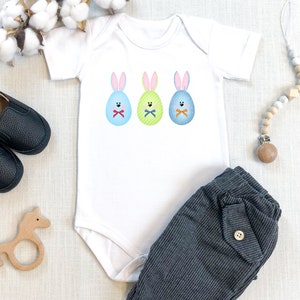 May include: A white baby onesie with three Easter bunny eggs on the front. The eggs are blue, green, and yellow with bunny ears and faces. The onesie is paired with gray striped pants.