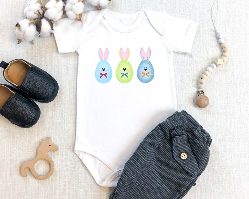 May include: A white baby onesie with three Easter bunny eggs on the front. The eggs are blue, green, and yellow with bunny ears and faces. The onesie is paired with gray striped pants.