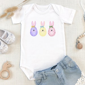 May include: A white baby onesie with a graphic of three Easter eggs with bunny ears and faces. The eggs are purple, yellow, and pink.