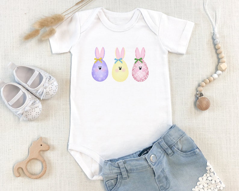 May include: A white baby onesie with a graphic of three Easter eggs with bunny ears and faces. The eggs are purple, yellow, and pink.