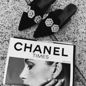 May include: A pair of black velvet pointed-toe mules with large rhinestone embellishments. The shoes are sitting on top of a Chanel Times magazine.