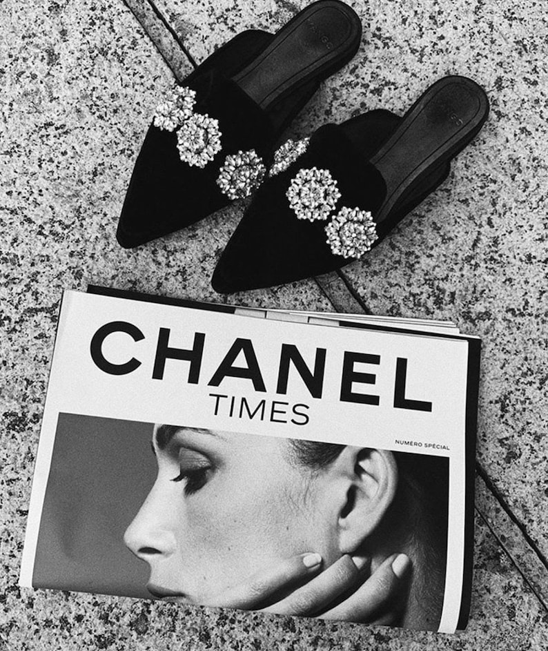 May include: A pair of black velvet pointed-toe mules with large rhinestone embellishments. The shoes are sitting on top of a Chanel Times magazine.
